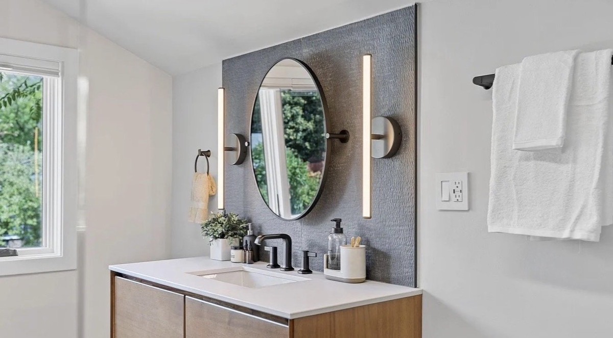 Bathroom with an oval mirror, linear sconces, gray tile wall and walnut vanity