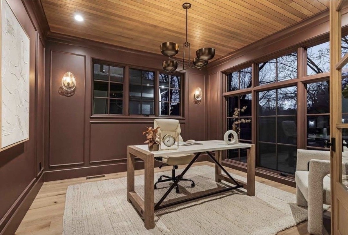 Moody home office with mauve paneled walls, a stained wood ceiling, brass chandelier and trestle desk