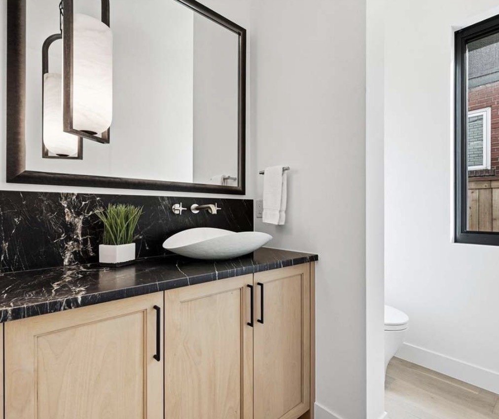 Powder room with a black marble counter, white vessel sink, oak cabinet and black framed mirror